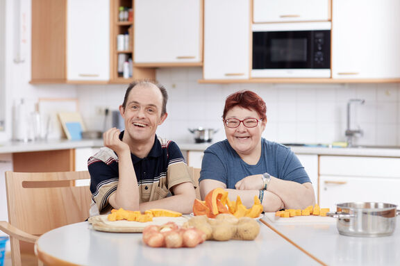 Zwei Menschen mit Behinderung sitzen in einer Küche und schneiden Obst und Gemüse
