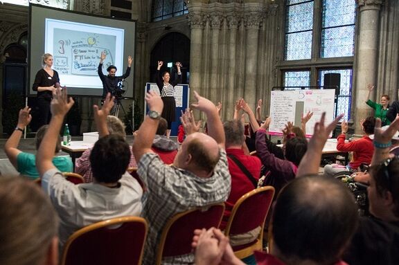 Das Publikum im Rathaus übt den Gehörlosen-Applaus. Es hebt die Arme und winkt mit den Händen. | www.mediengarten.eu Das Publikum im Rathaus übt den Gehörlosen-Applaus. Es hebt die Arme und winkt mit den Händen.